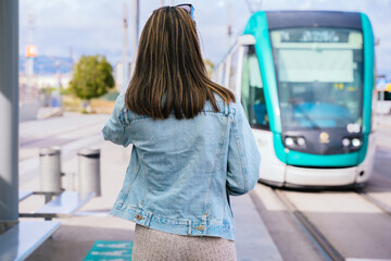 Woman in a denim jacket standing at a tram stop, facing an approaching tram. She waits for public transportation in a modern urban setting, ready for her commute or journey.