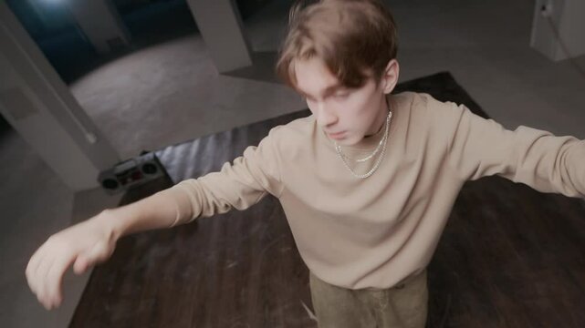 Handheld high angle shot of creative Caucasian male zoomer in streetwear and hip hop chain improvising top rock breaking moves while dancing to music on boombox indoors