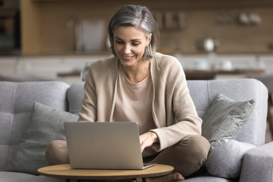 Cozy remote job. Mid aged woman self employed entrepreneur sit on sofa at home office workplace do accounting work for remote client create content for business blog on notebook research market trends