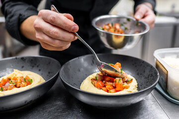 A chef expertly plating hummus topped with fresh tomato salsa in a modern kitchen. Fresh ingredients are being carefully arranged in elegant black bowls.