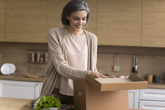 Home food delivery. Smiling middle aged woman housewife satisfied grocery store customer unpacking postal parcel box with fresh natural organic healthy food products ordered online at domestic kitchen