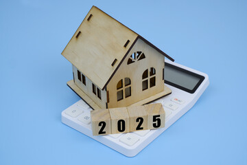 House model and wooden cubes with numbers 2025 on white calculator on blue background. Cost of real...