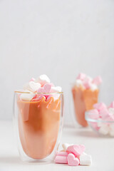 Festive hot chocolate or cocoa drink decorated with pink heart shaped marshmallow dessert snack in glass mug or cup served on white wooden table with ingredients on valentines day holiday celebration