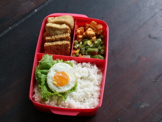 Balanced nutritious food consisting rice, egg, tofu, tempeh, and vegetables, served on lunch box, suitable for students health