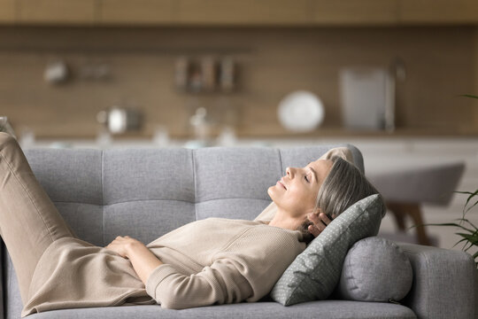 Daily nap for self care. Calm serene early senior age lady take break to relax at day time lying on back on comfortable couch dream sleep rest with closed eyes breath fresh conditioned air. Copy space