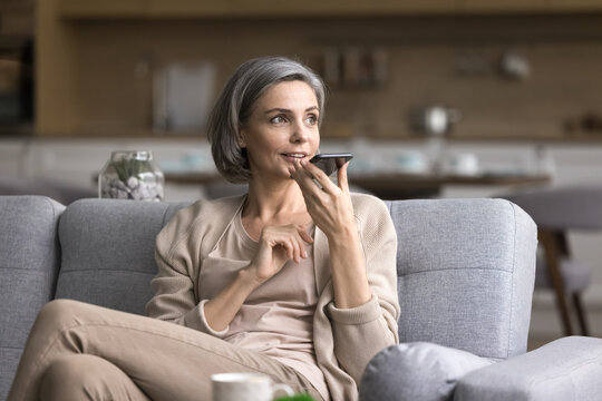 Relaxed communication. Inspired elegant mature woman sit on couch speak on phone holding device close to mouth use speakerphone to dictate vocal command to ai assistant record voicemail audio message