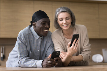 Digital communication in marriage. Bonding friendly middle aged multiethnic couple of spouses share information between two smartphones at kitchen send receive photo video files using wifi connection