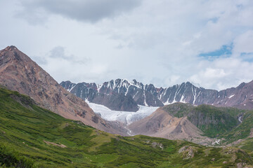 Obraz premium Scenic landscape of green alpine valley. Lush flora on hills and rocks with view to big glacier and large snow mountain range far away under gray cloudy sky. Awesome mountains in changeable weather.