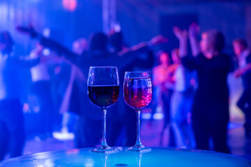Two wine glasses on table amidst lively party atmosphere with people dancing and vibrant lighting.