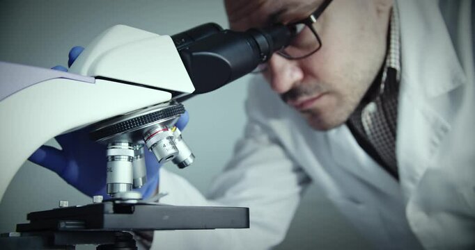 Researcher, wearing lab coat, is meticulously examining various slides under highpowered microscope
