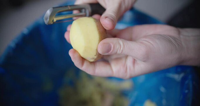 Person carefully peels fresh potato, discarding the skin properly into blue trash bin nearby