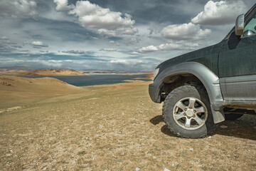 An off-road vehicle with mud tires stands in the steppe against the background of the sky and the lake. A tourist car on non-hydrogen tires with a view of the far lake. © ANDREY