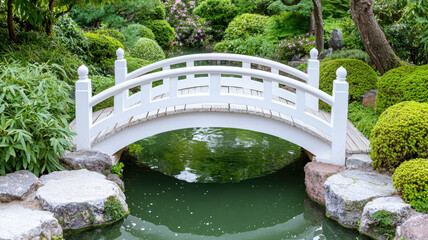 A serene garden scene featuring a white bridge over a tranquil pond, surrounded by lush greenery and neatly shaped bushes.