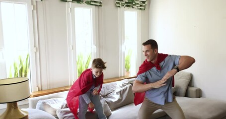 Happy active dad and excited playful son kid wearing superhero handmade cape, dancing at home, having fun, playing active game. Strong daddy lifting and spinning child