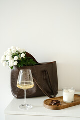 A chic composition featuring a stylish designer bag with fresh flowers , a beautiful glass of wine, chocolote, candle on wooden tray.