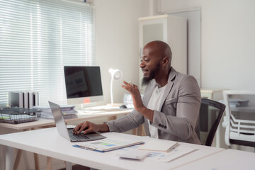 African american businessman waving to someone during a video call while working on his laptop in a modern office, surrounded by paperwork and technology