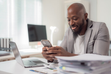 Obraz premium Happy african american manager texting on mobile phone while working at modern office with laptop and stack of documents, enjoying online communication or using business app