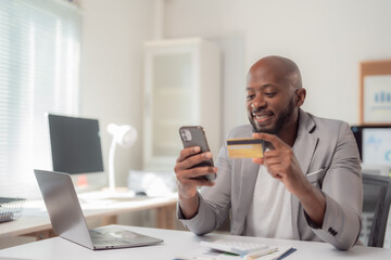 Smiling African American businessman holding a credit card while using a smartphone for seamless online shopping at his modern workplace, enjoying the convenience of digital transactions