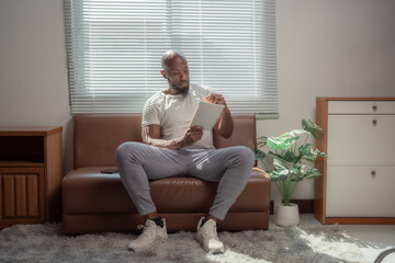 Concentrating on notes while reading from a tablet, an African American man relaxes on a cozy sofa in a comfortable living room, enjoying leisure time at home