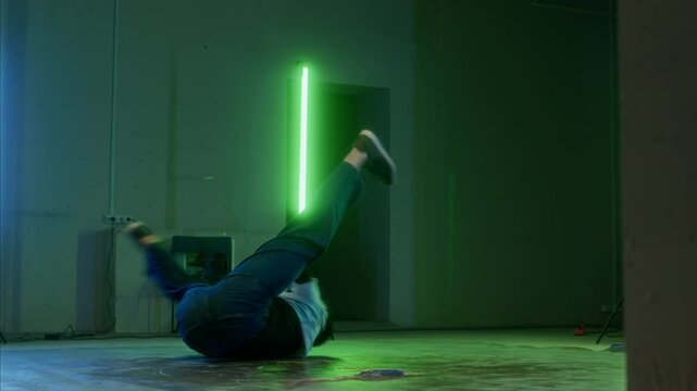 Slowmo of young talented Caucasian breakdancer spinning his body performing breathtaking acrobatic movements under green and blue lamps indoors