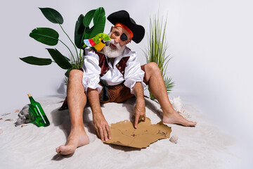 Old bearded pirate with parrot examines treasure map on sandy island beach surrounded by tropical...