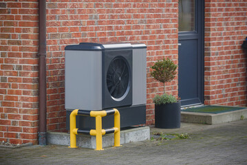 Modern heat pump system installed outside a red-brick house, protected by a yellow and black safety barrier. Sustainable heating solutions for building. Air source heat pump beside residential house. 
