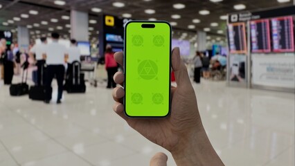 GREEN SCREEN CHROMA KEY A hand holding a smartphone with a green screen in an airport terminal. Blurred travelers and modern architecture in the background
