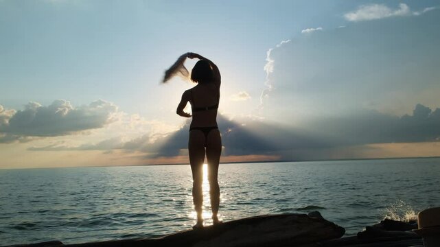 Grace silhouette of a beautiful bikini woman dancing by the sea at sunset. Young pareo girl standing on the beach with setting sun on the background.