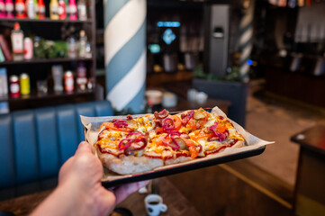 A close-up of a freshly baked pizza topped with cheese, pepperoni, pickles, and onions, served on a black tray in a cozy restaurant setting. The ambiance is warm and inviting.