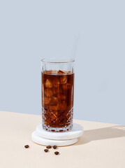 Cold brew coffee in a tall glass with ice and straw on a marble stand on a yellow-blue background