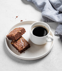 Pieces of chocolate brownie with nuts on a plate with a cup of espresso coffee on a light background