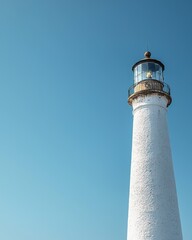 Fototapeta premium A tall white lighthouse stands against a clear blue sky, symbolizing guidance and safety for ships navigating the waters.