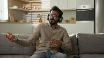 Happy relaxed Arabian man homeowner listen music in headphones enjoy rhythm song pretend playing drums on home sofa cheerful Indian guy listening musical track in earphones energetic dance in kitchen