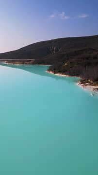 A turquoise tailings pond near a cement factory, surrounded by rugged hills. The vivid water contrasts with the barren land, creating a striking yet industrial scene of environmental impact