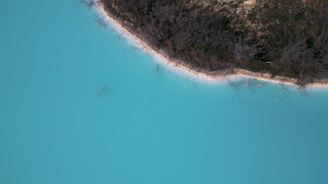 A turquoise tailings pond near a cement factory, surrounded by rugged hills. The vivid water contrasts with the barren land, creating a striking yet industrial scene of environmental impact