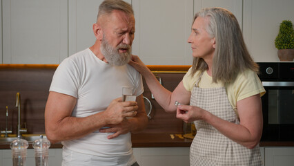 Sick unhealthy old man in kitchen feeling pain in chest heart attack heartache bad health cardiac medical problem Caucasian family caring mature woman wife help husband give remedy tablet drink pill