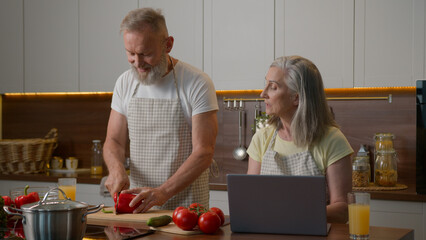 Caucasian mature love family in home kitchen middle-aged woman wife shopping online food delivery buy products internet store with laptop senior adult man husband cut fresh vegetables cooking dinner