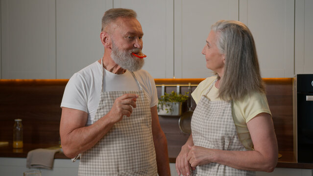 Caucasian couple middle-aged old family woman feeding beloved man eat piece of pepper red paprika slice healthy vegetarian food taste fresh vegetables eating wife and husband cook together at kitchen