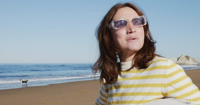 Mujer con cabello rojo feliz se pone las gafas de sol, disfrutando en la playa los primeros d&iacute;as de primavera, en un d&iacute;a soleado, se pone las gafas de sol, frente al mar. Plano corto