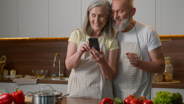 Happy senior old couple love bonding family adults woman man browsing smartphone online order delivery fresh food search recipe breakfast on mobile phone retired wife husband talk cook at home kitchen