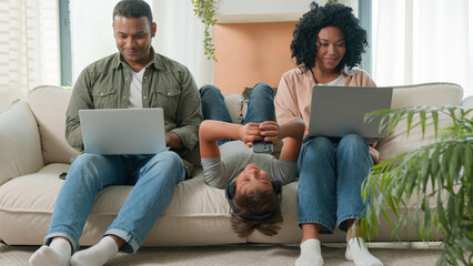 Addicted family use gadgets devices little kid child son boy lying upside down on sofa play video game on mobile phone smiling African American parents working laptops different gen using modern tech