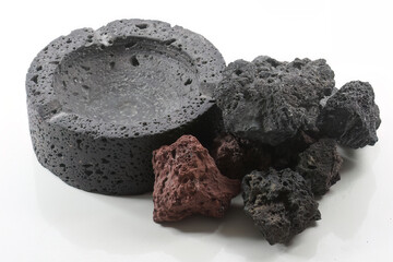 Black ashtray of volcanic stone and pieces of raw pumice-stone on white.