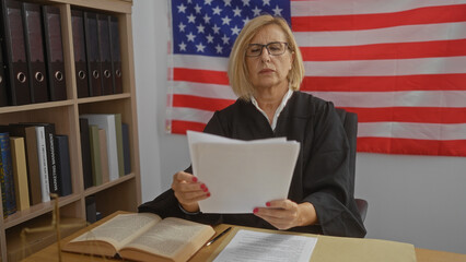 Woman judge in a courtroom reading documents with american flag, books, and binders on shelves in...