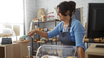 Woman working in a bakery shop, serving gluten-free pastries with tongs in a bright indoor setting...