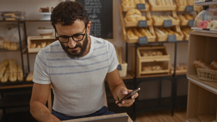 Young man with beard and glasses using phone in bakery shop with bread in background