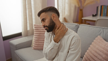Young man with a beard touching his neck in pain while sitting in a living room, wearing a light...