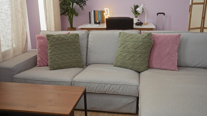 Elegant living room with a gray sofa adorned with green and pink pillows, wooden coffee table, potted plants, and a study desk in the background illuminated by a neon light.