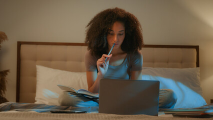 African American woman girl student studying with laptop writing notes homework preparing for exams on bed in evening night home bedroom online course learning distant education e-learning on computer