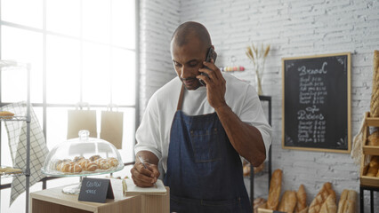 Young man in a bakery wearing an apron talking on phone while taking notes near pastries and bread display