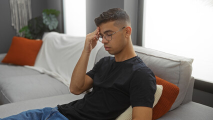 Obraz premium Young man looking worried at home sitting on couch in modern living room with potted plants and red cushions wearing glasses and casual outfit.
